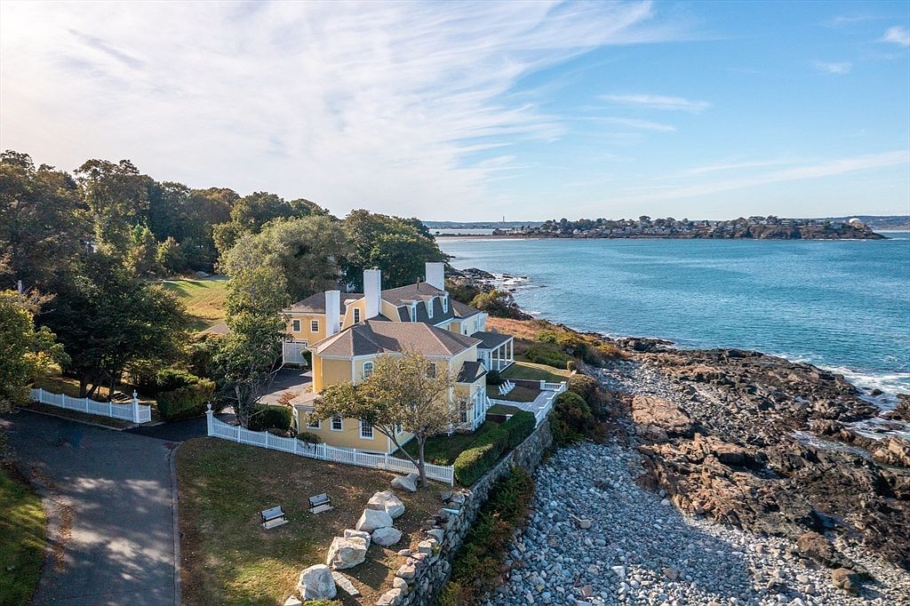Late-afternoon aerial of 66 Ocean Street, the yellow estate behind a white perimeter fence with lawns running to the rocky shoreline and open ocean beyond