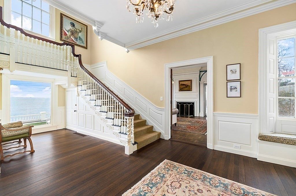 Two-story stair hall with a carved white balustrade, mahogany handrail, a tall window framing the ocean, and a gallery landing above
