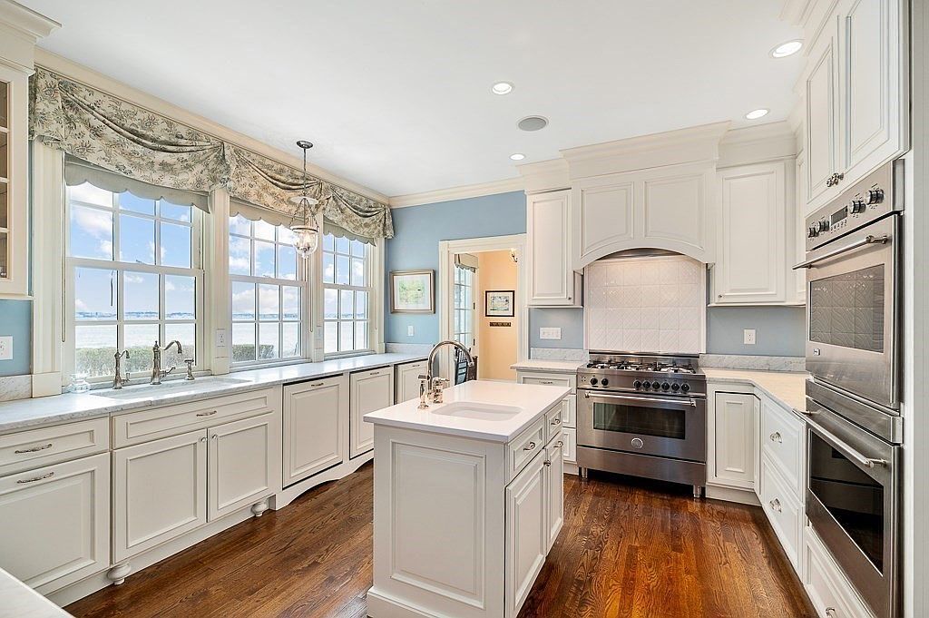 Chef's kitchen with white raised-panel cabinetry, marble counters, a professional range, double wall ovens, and windows over the water above the sink