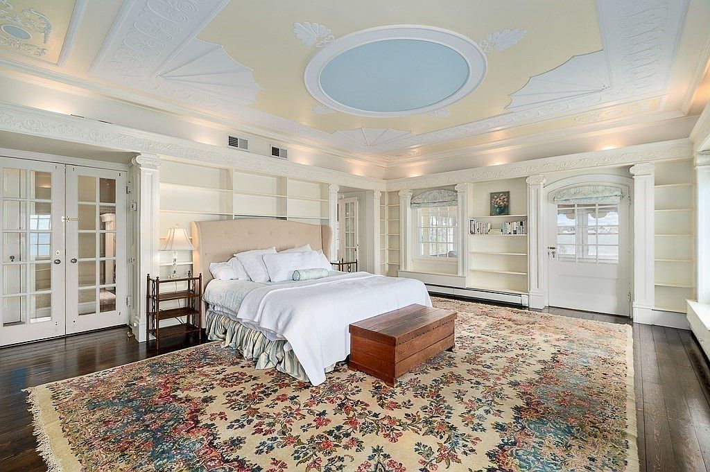 Primary bedroom with paneled walls, a fireplace, an ornate painted ceiling with a pale blue oval medallion, and French doors framing the water