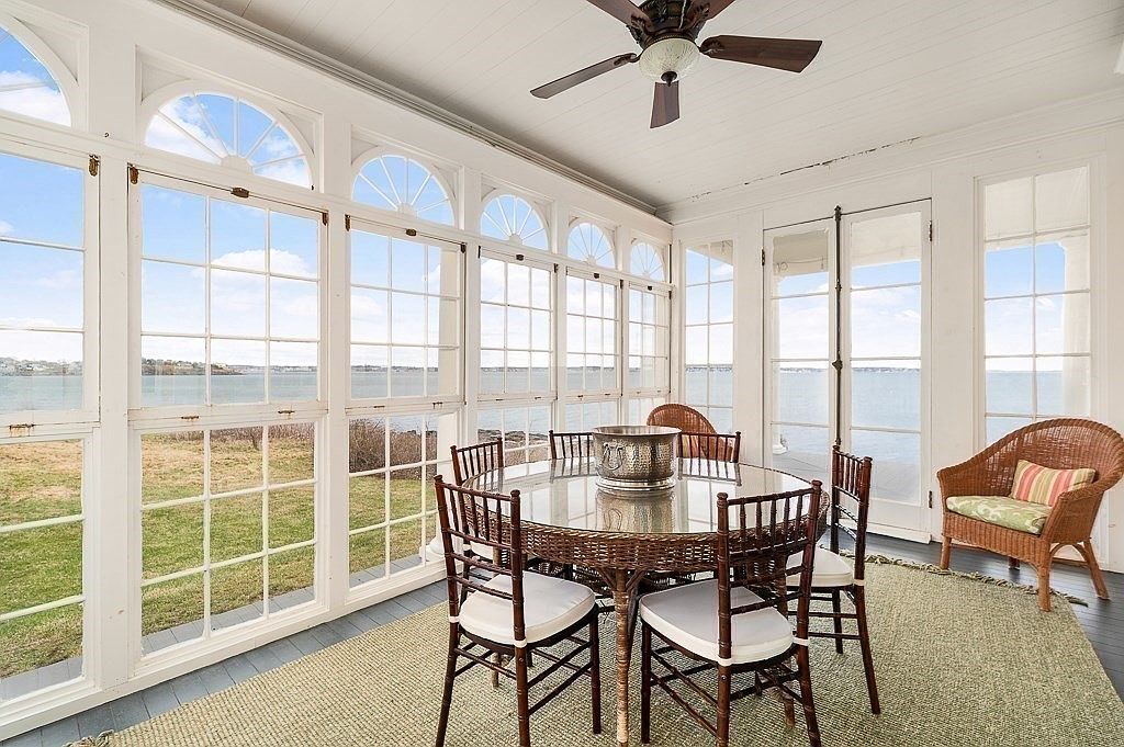 Glassed sunroom wrapped in arched transom windows on three sides, looking out over the lawn to the water, with wicker furniture and a glass table