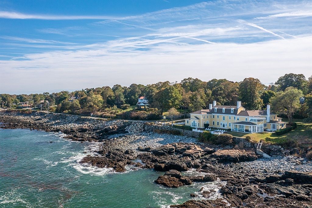 Aerial view of 66 Ocean Street, a yellow 1890 oceanfront mansion perched above the rocky Nahant shoreline with surf breaking on the rocks and open water beyond